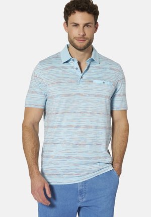 Mann trägt ein hellblau gemustertes kurzärmeliges Poloshirt mit Kragen und vorderen Knöpfen, kombiniert mit blauen Jeans, Hände entspannt.
