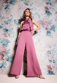 Vluchtige roze jumpsuit met wijde benen en een halternek met een strikaccent. Gladde stof tegen een bloemenpatroon achtergrond.