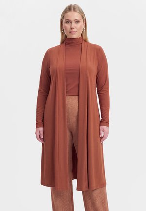 Vrouw die een roestkleurige coltrui draagt, bijpassende lange open cardigan en broek met visgraatpatroon, staand tegen een effen achtergrond.