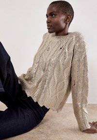 Maglione lavorato a maglia in oro con un design a treccia testurizzata e orlo frangiato. Abbinato a denim scuro, che mostra una vestibilità aderente in vita.