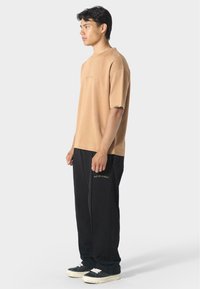 Béžová košile s krátkým rukávem a nenápadným logem, doplněná černými kalhotami s výšivkou, a černými teniskami s bílými tkaničkami.