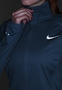 Personne portant une veste zippée bleu foncé avec le logo Nike, ajustant le col d'une main, visible du cou à la taille.