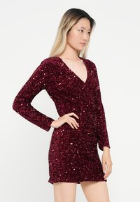 Burgundijas sekmētu kleita ar garām piedurknēm, dziļu V-veida kakla izgriezumu un pieguļošu vidukli. Tekstūra ir gluda un spīdīga, ar īsu hemline.