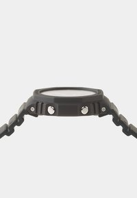 Montre en plastique grise avec un cadran rond, comportant deux boutons latéraux, un bracelet strié et des accents métalliques sur les boutons.