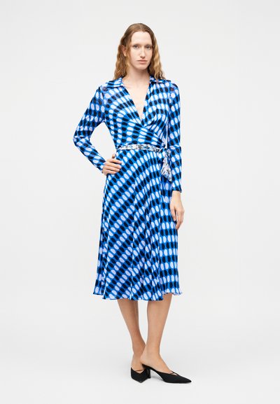 Γυναίκα με μπλε φόρεμα τύπου wrap με κυκλικά μοτίβα, μακριά μανίκια, μέση με ζώνη και μαύρα παπούτσια με μύτη και χαμηλό τακούνι, στέκεται με το χέρι στη μέση.