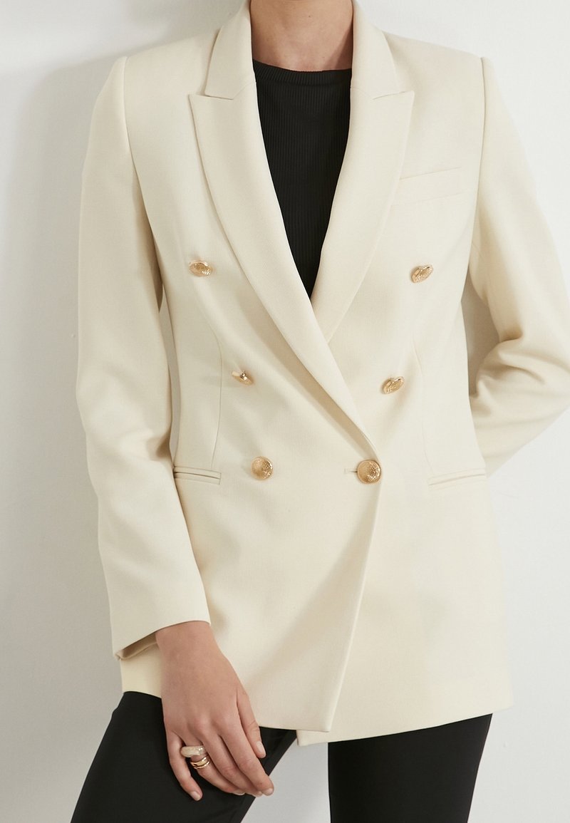Double-breasted blazer in cream fabric, featuring gold-tone buttons, notched lapels, and two side pockets. Textured material with structured fit.