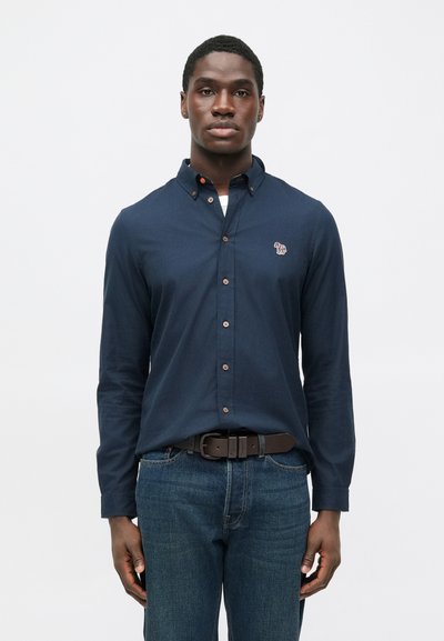 Camicia blu navy a maniche lunghe con bottoni e colletto, con logo ricamato sul petto, abbinata a jeans di denim blu e cintura marrone.