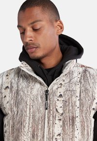 Gestepptes Weste mit einem weißen und braunen Birkenbaum-Muster, mit einem hohen Kragen und einem Reißverschluss auf der Vorderseite. Über einem schwarzen Hoodie getragen.