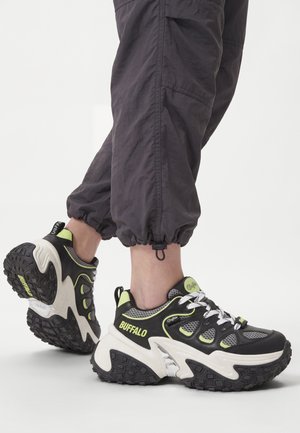 Persona che indossa sneakers chunky Buffalo nere e verde neon con pantaloni neri e grigi risvoltati su uno sfondo chiaro e uniforme.