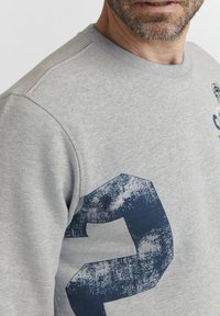 Grauer Pullover mit Rundhalsausschnitt, ausgestattet mit einem strukturierten blauen Design der Zahl 2 und kontrastierenden orangefarbenen Nähdetails an der Schulternaht.