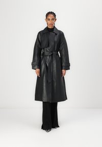 Čierny kožený trench coat so širokým golierom, opaskom v páse a veľkými rukávmi. Nosený cez priliehavé čierne nohavice, s elegantnými, lesklými topánkami.