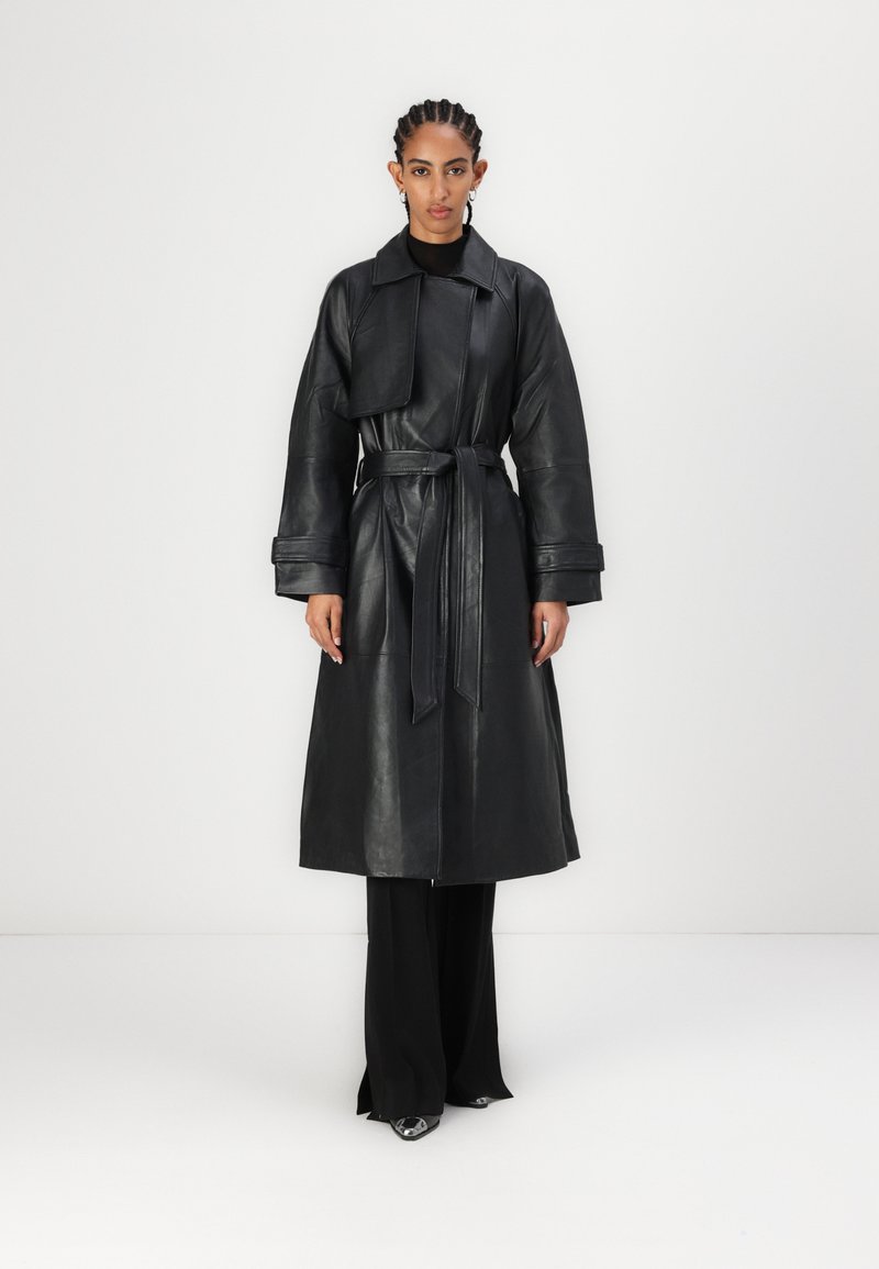 Čierny kožený trench coat so širokým golierom, opaskom v páse a veľkými rukávmi. Nosený cez priliehavé čierne nohavice, s elegantnými, lesklými topánkami.