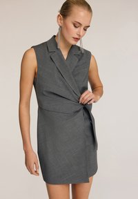 Abito grigio senza maniche con rever a scollatura a V, design avvolgente e dettaglio con fiocco laterale. Realizzato in tessuto testurizzato con accenti di cucitura a contrasto.