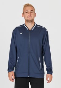 Marineblå atletisk jakke med hvid piping, fuld lynlås og sidelommer. Fremstillet af glat, letvægtsmateriale med et klassisk kravedesign.