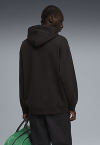 Sudadera negra con capucha y textura suave; cuenta con un bolsillo tipo canguro y puños de canalé, combinada con un bolso verde y pantalones oscuros.
