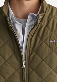 Quilted olive green jacket with a zippered front, featuring a tonal collar and logo embroidery. Layered over a light blue striped shirt.