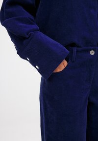 Tamsiai mėlynos spalvos kordūrojo audinys puošia marškinėlius su tekstūruotu dizainu ir segamomis rankovėmis, derinami su atitinkamomis kelnėmis, turinčiomis priekinį kišenę.