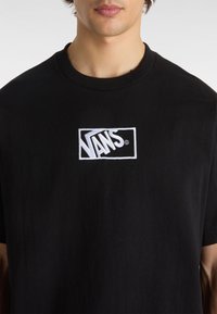 Camiseta de algodón negra con un logo de Vans bordado en blanco en un diseño rectangular en el pecho. Cuello redondo y mangas cortas.