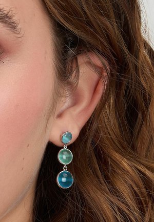 Primo piano dell'orecchio con orecchino pendente che presenta tre pietre rotonde con sfumature blu-verdi incastonate in argento. Capelli castano chiaro e mossi visibili.