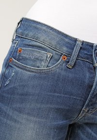 Tätt sittande blå denimjeans med en klassisk femficksdesign, med kopparfärgad hårdvara, blekta områden och en subtil slitna struktur.