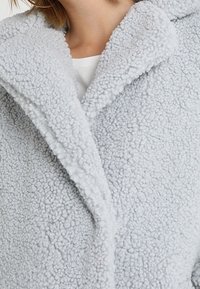 Cappotto in pile grigio chiaro con una superficie texture e pelosa, dotato di un ampio colletto e una chiusura con bottoni per una vestibilità rilassata.