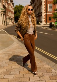 Brązowe sztruksowe spodnie w rozszerzanym kroju, szary golf, żakiet w zwierzęcy wzór oraz szare sneakersy. W okularach przeciwsłonecznych, idąc po brukowanej chodniku.