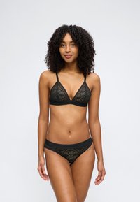 Femme aux cheveux bouclés portant un soutien-gorge en dentelle noire et une culotte assortie, debout devant un fond gris clair uni.