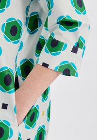 Camicia a fantasia con forme geometriche verdi e blu su sfondo bianco. Maniche con polsini e dettaglio con bottone e tasca laterale.