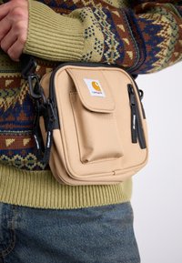 Beige stof skuldertaske med to sorte lynlåslommer, en frontklap og et logo-patch, båret over en mønstrede sweater.