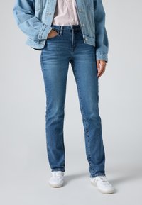 Dark blue denim jeans with a straight fit, mid-rise waist, and slight whiskering. Paired with a light blue denim jacket and white sneakers.