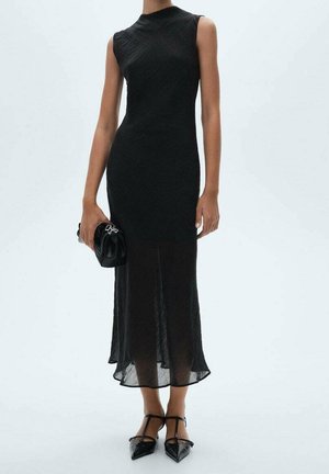 Kobieta w czarnej, bezrękawnikowej sukience midi, trzymająca czarną kopertówkę i mająca na nogach czarne szpilki z paskiem typu T i spiczastym noskiem.