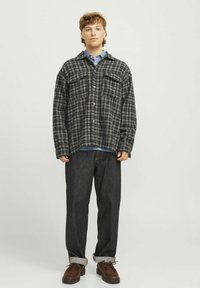 Sötétzöld és szürke kockás flaneling, két elülső zsebből, gombos, világoskék farmering fölött viselve, sötét farmernadrággal és barna cipővel.