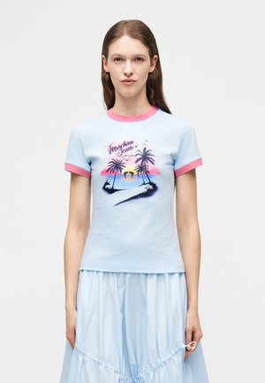 Mladá žena s dlhými hnedými vlasmi, oblečená v svetlomodrom tričku s ružovým lemovaním a vzorom tropického západu slnka s palmou.
