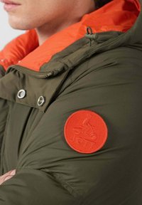 Chaqueta acolchada de color verde oliva con textura acolchada, que presenta un cuello interior naranja y un parche redondo de logo rojo en la manga.