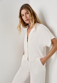 Blusa leggera a maniche corte di colore crema, con una vestibilità morbida, chiusura a bottoni e un leggero luccichio. Abbinata a pantaloni larghi coordinati.