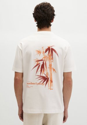 Man die een wit korte mouwen shirt draagt met oranje en rode bamboe afbeelding en tekst "Bamboo Lounge" op de rug, staand tegen een effen achtergrond.