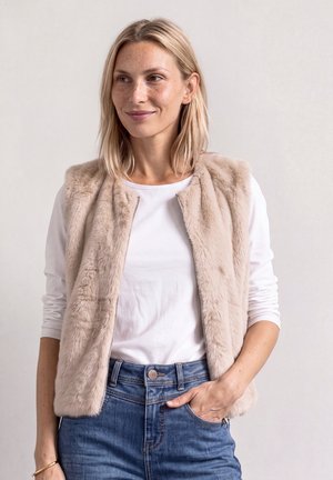 Blonde vrouw met een beige nep bontvest over een wit shirt met lange mouwen en een blauwe spijkerbroek, met één hand in de zak, licht glimlachend.