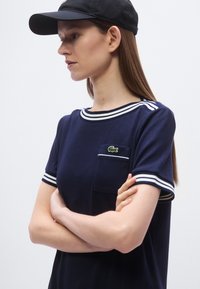 T-shirt marine côtelé avec des rayures athlétiques blanches sur le col et les manches, doté d'un petit logo vert et d'une poche poitrine.