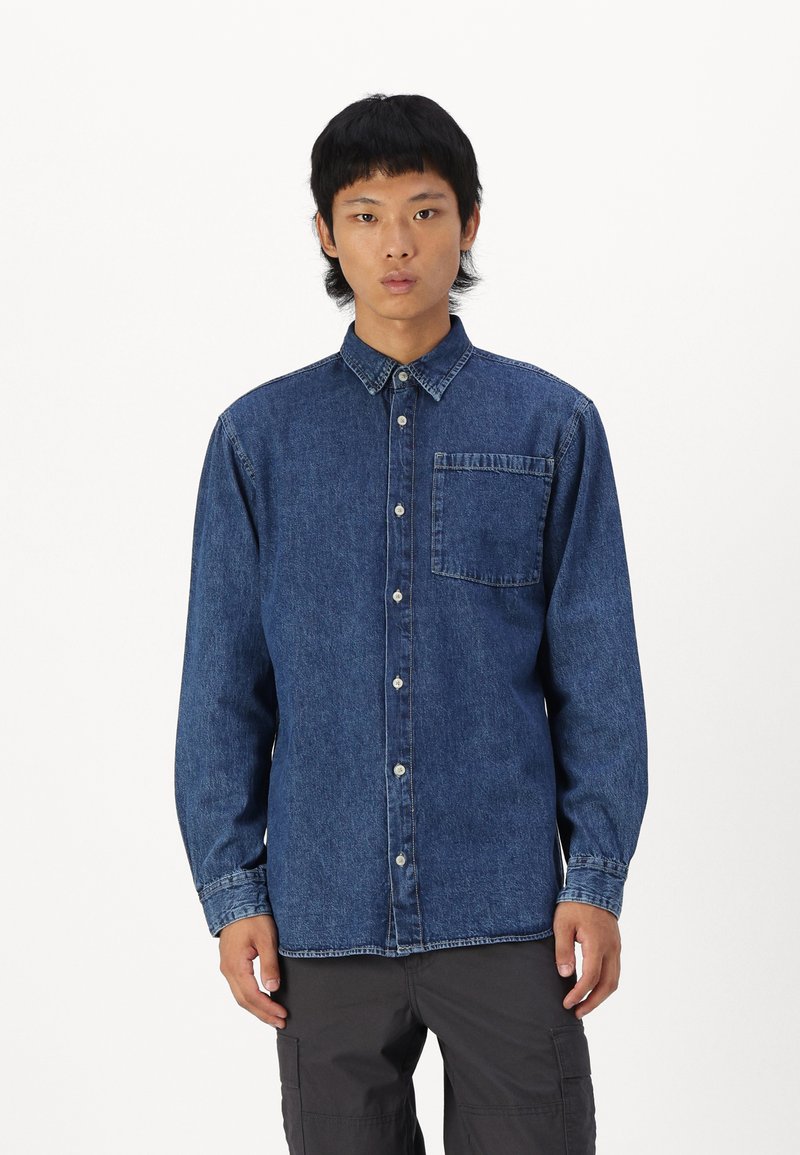 Camisa de mezclilla en azul oscuro con mangas largas, cierre de botones en la parte frontal y un solo bolsillo en el pecho. Presenta un cuello clásico y un corte relajado.