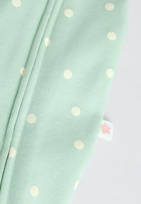 Világoszöld anyag fehér pöttyökkel, látható varrás és egy kis fehér címke rózsaszín virágdísszel világos háttéren.