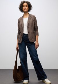 Blazer lungo marrone a maniche lunghe su camicia bianca, abbinato a jeans larghi blu scuro e sneakers bianche. Una borsa tote marrone completa l'outfit.