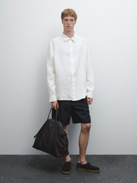 Witte linnen overhemd met knopen, zwarte shorts, donkerbruine leren tas en donkere schoenen met lichte zolen. Eenvoudig ontwerp en neutrale kleuren.