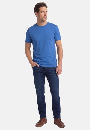 Mężczyzna w niebieskiej koszulce z krótkim rękawem, ciemnoniebieskich dżinsach i brązowych trampkach, stojący z jedną ręką w kieszeni, patrzący na bok.