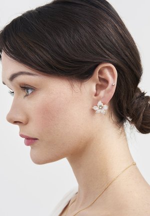 Woman with dark hair in a low bun wearing a white flower-shaped earring and a delicate gold necklace, facing left against a plain background.