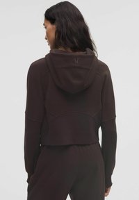 Nő sötétbarna cropped kapucnis felsőben és ahhoz illő nadrágban, hátulról nézve, sima világos háttér előtt.