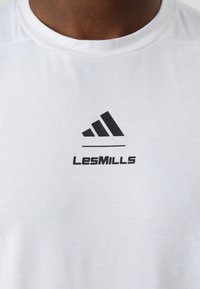 Bílý sportovní tričko s kulatým výstřihem, na přední straně se černým textem "LesMILLS" a černým logem se třemi pruhy. Měkká textura.