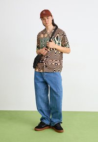 Chemise à manches courtes à motifs abstraits multicolores, associée à un jean en denim bleu ample et des baskets noires. Casquette marron avec détail de logo.