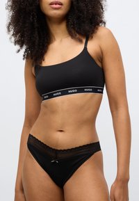 Czarny bezszwowy biustonosz typu bralette z cienkimi ramiączkami i białą opaską z logo; zestawiony z pasującymi czarnymi stringami ozdobionymi koronką i kokardką.