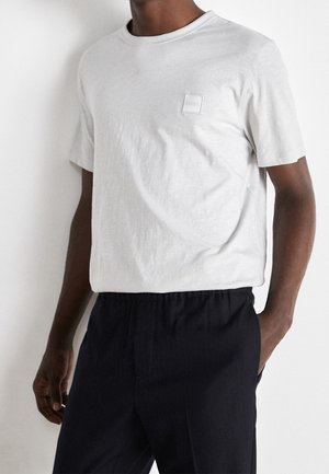 Osoba w jednolitym białym T-shircie z krótkim rękawem, z małym kwadratowym logo na piersi, ubrana w czarne spodnie, z jedną ręką w kieszeni.