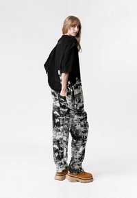 Joven de pie de lado vistiendo una camisa negra de gran tamaño, pantalones de patrón blanco y negro con etiquetas, y zapatos tipo plataforma gruesos color canela sobre un fondo liso.